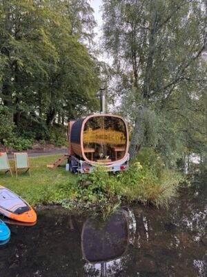 Vista posteriore con vetrata panoramica della sauna mobile TimberIN Nordic Square su rimorchio accanto a un lago