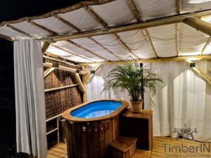 TimberIN Nordic Duo compatta in vetroresina con acqua blu, rivestimento in legno termotrattato e scaletta in legno sotto un gazebo bianco con una grande pianta in vaso