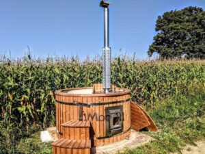 TimberIN Nordic Flex su un prato davanti a un campo di mais, con scaletta in legno, frontale in acciaio inox della stufa, alto camino e coperchio in legno