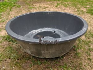 Vasca grigia in vetroresina vuota per la TimberIN Nordic InGround Comfort su un prato, con panca rotonda visibile