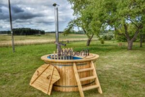 TimberIN Nordic Flex aperta in un parco, vista di tre quarti, con vasca interna blu, scaletta a destra, due coperchi in legno appoggiati e alto camino