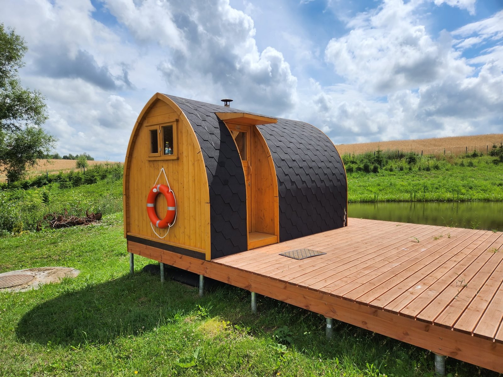 Casetta da giardino in legno a forma di igloo con tetto curvo nero, porta e finestra, su una terrazza in legno vicino a un lago