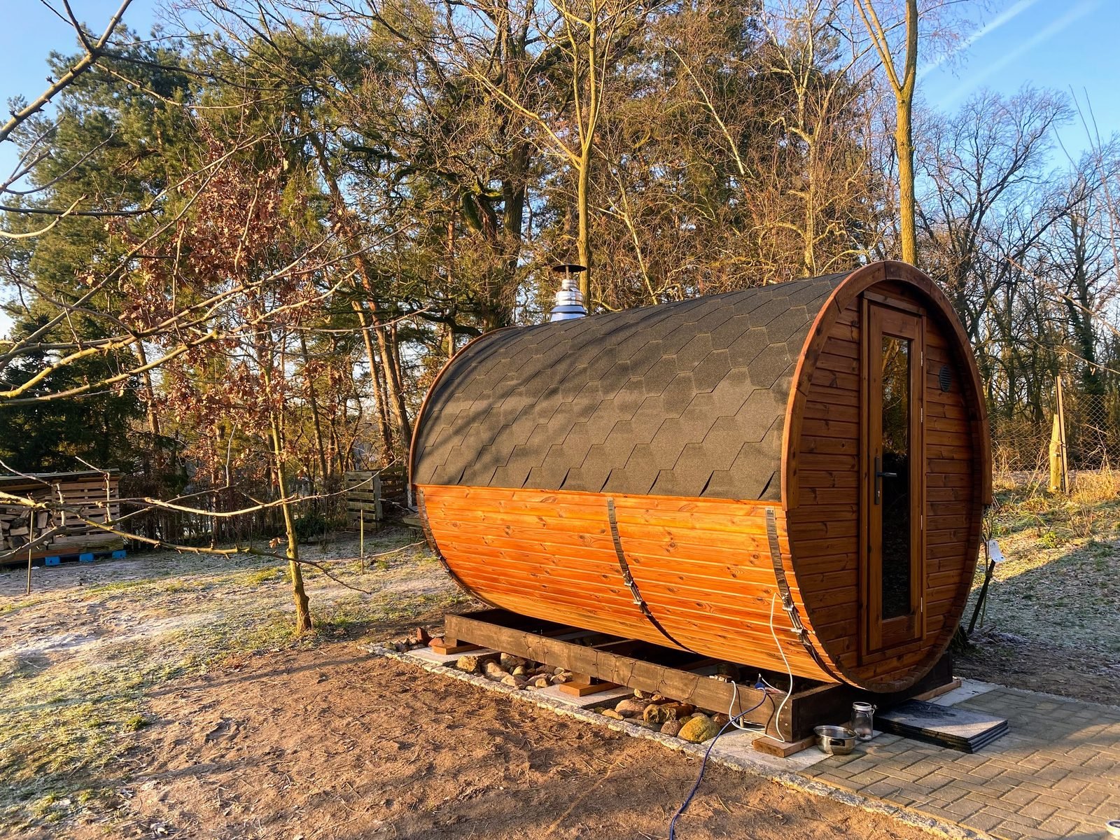 Piccola sauna a botte in legno con tetto nero e porta in vetro, su una base sotto gli alberi del giardino