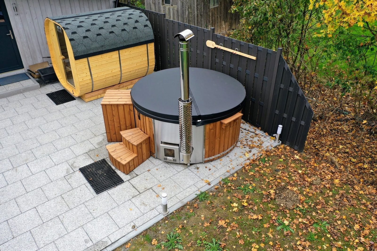Sauna a botte e vasca in legno con stufa a legna esterna, affiancate su un patio in giardino