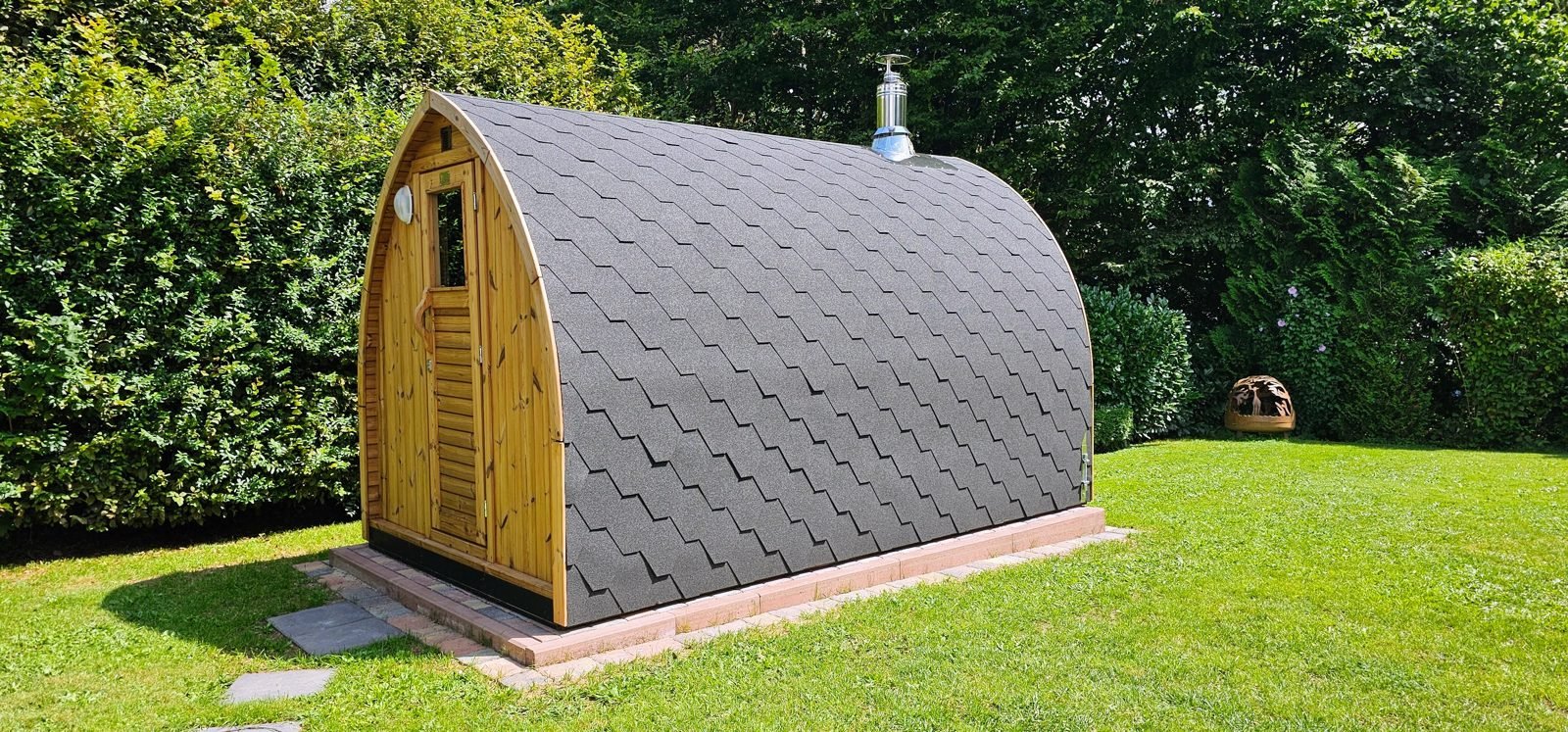 Sauna da esterno in legno con tetto curvo a scandole e canna fumaria su un prato in giardino