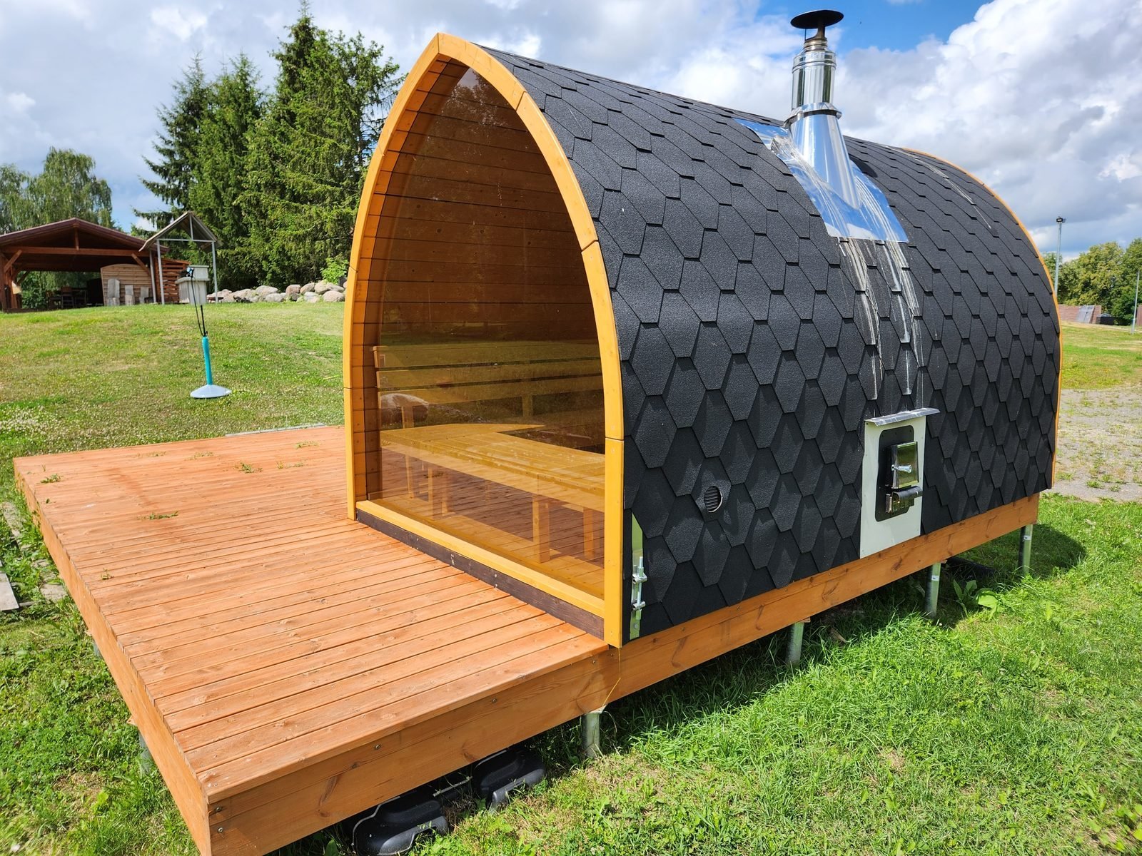 Sauna da esterno in legno con grande vetrata frontale, tetto a scandole nere e camino, su una terrazza in legno in un prato