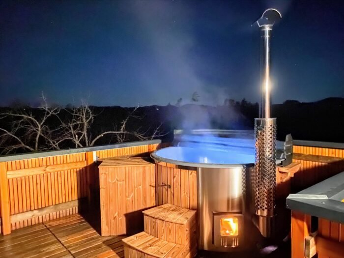Vasca idromassaggio da esterno in legno con stufa a legna e camino su una terrazza in legno, di sera