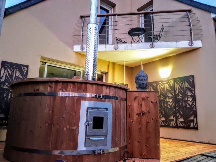 Vasca idromassaggio in legno con stufa a legna e camino su una terrazza illuminata di sera