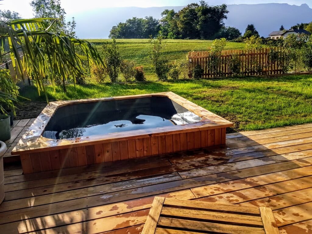 Vasca idromassaggio quadrata con vasca interna scura incassata in una terrazza in legno, con vista sul prato