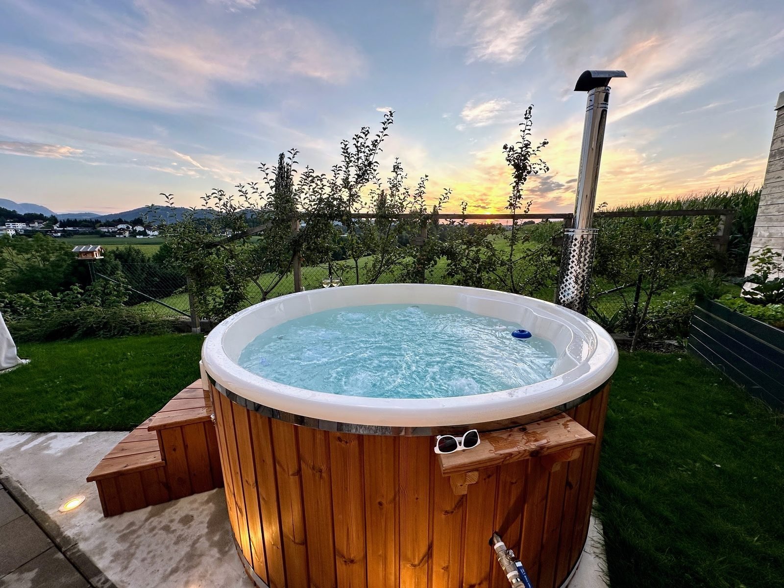 Vasca idromassaggio rotonda con vasca interna chiara in vetroresina, acqua con bolle e rivestimento in legno su una terrazza al tramonto