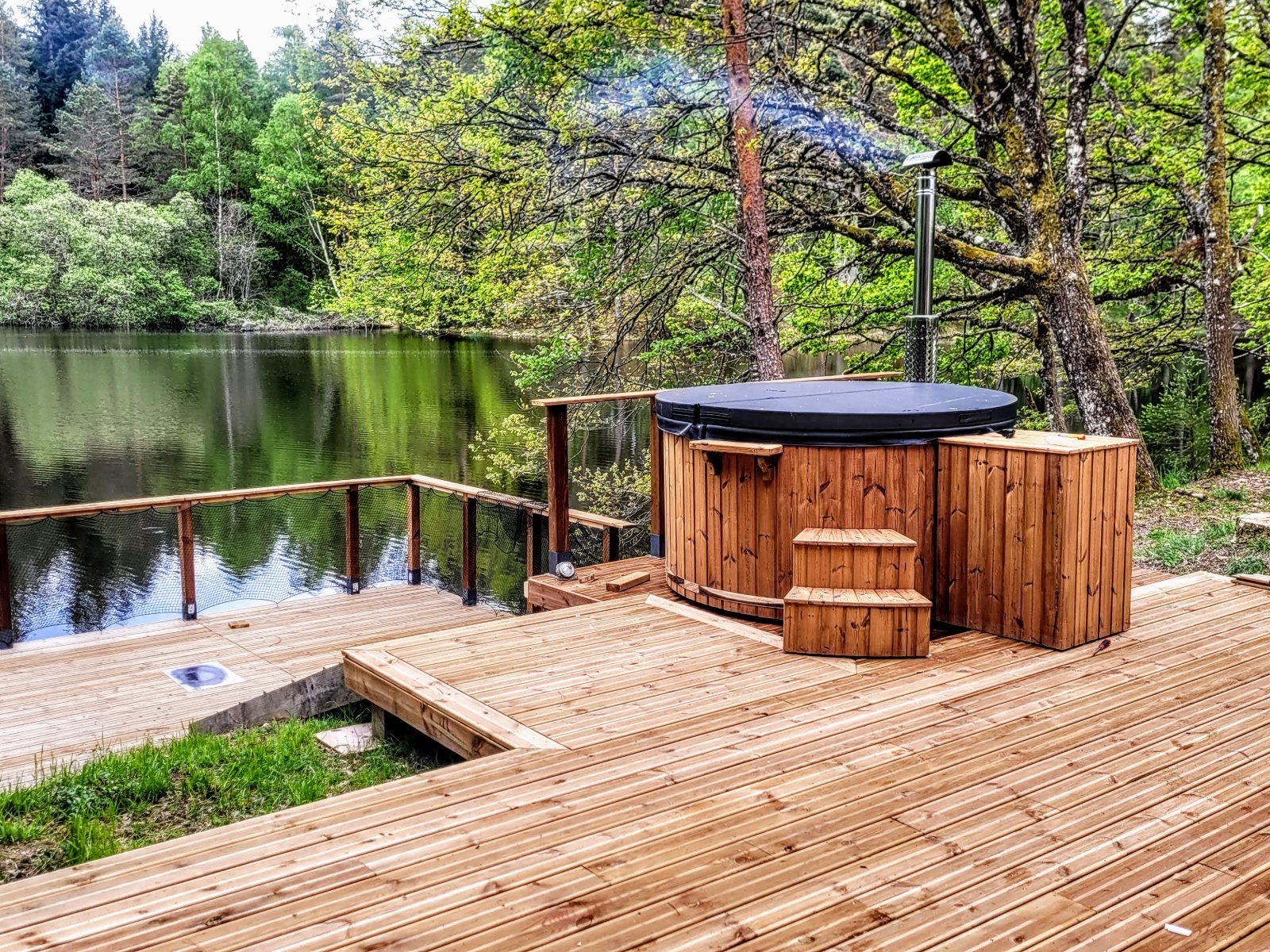 Vasca rotonda in legno con coperchio e scaletta su una terrazza in legno vicino a un lago nel bosco