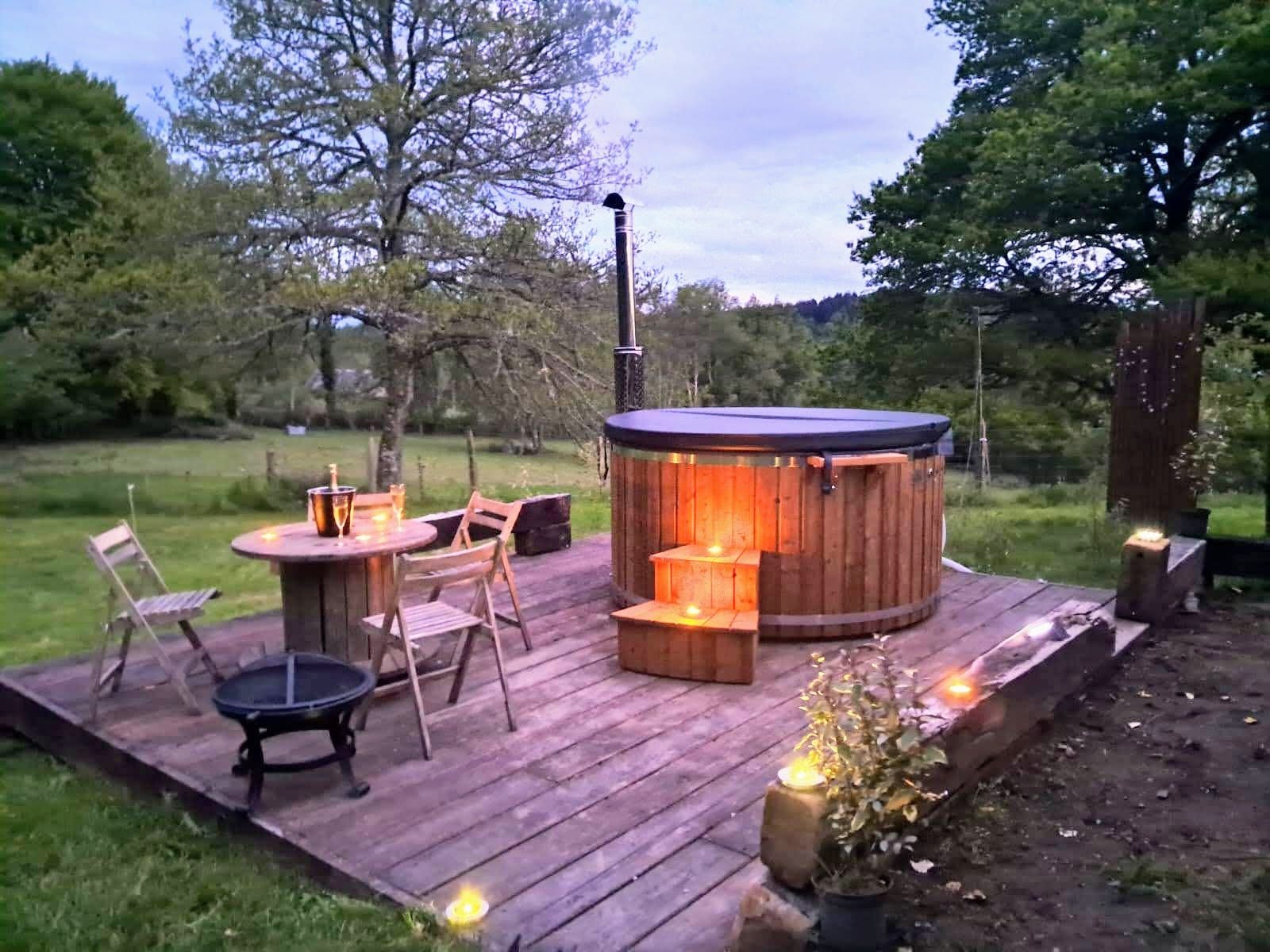Hot tub rotondo in legno con luce calda su una terrazza in legno in giardino al crepuscolo, con tavolo e sedie